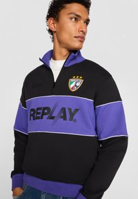 Sort og lilla sweatshirt med lynlås krave, som har et stort "REPLAY" logo og et våbenskjold med tre stjerner. Blødt stof.
