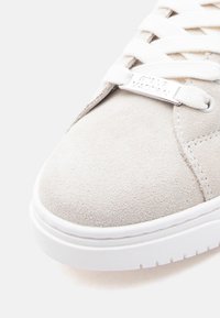 Κοντινή εικόνα της μύτης ενός μπεζ σουέντ αθλητικού παπουτσιού με λευκά κορδόνια και μεταλλική ετικέτα με χαραγμένο το "Steve Madden" στα κορδόνια.