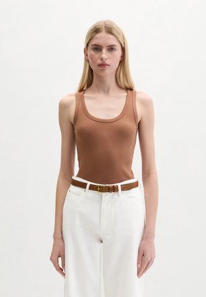 Femme aux longs cheveux blonds portant un haut marron sans manches rentré dans un pantalon blanc taille haute avec une ceinture marron, debout devant un fond uni.