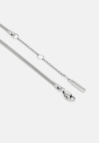 Silberne Kette mit einem runden Glieddesign, ausgestattet mit einem Clip und einem rechteckigen Tag, der mit "Calvin Klein" graviert ist. Glatte Metalloberfläche. 