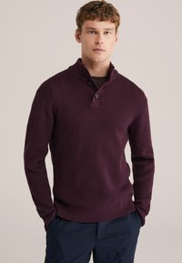 Bordeaux gebreide pullover met een hoge kraag, twee-knopen sluiting, geribde manchetten en een gestructureerd oppervlak. Gecombineerd met marineblauwe broek.