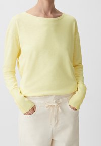 Gul långärmad tröja med vid rundhalsning och ribbade ärmmuddar, i kombination med ljusa byxor med dragsko. Mjuk textur, avslappnad passform.