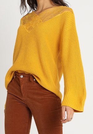 Frau trägt senfgelben Strickpullover mit Spitzen-Ausschnitt kombiniert mit rostfarbenen Cordhosen, Hand entspannt an der Seite.