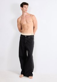 Schwarze, weit geschnittene Jeans mit einem hellen Bund. Der Stoff ist Denim mit einer glatten Textur. Das Model steht barfuß mit den Armen hinter dem Rücken.