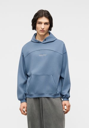 Jeune personne portant un sweat à capuche bleu surdimensionné avec une poche frontale et un pantalon gris ample, debout devant un arrière-plan uni.