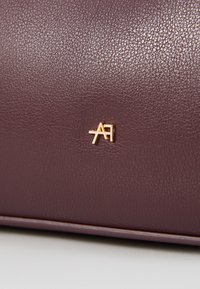Sac en simili cuir bourgogne avec une surface texturée et un emblème doré présentant les initiales "A" et "F". Bords lisses et éclat subtil.