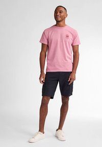 Ljusrosa bomullst-shirt med en liten grafik på bröstet, kombinerad med mörka cargobyxor och krämfärgade sneakers, ståendes mot en neutral bakgrund.