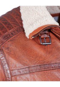 Bruine leren jas met een schapenvacht kraag, met een gestructureerd oppervlak, metalen rits en decoratieve stiksels over de schouders.