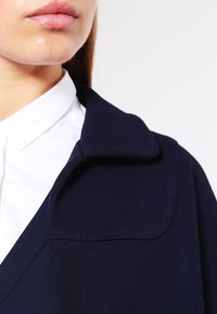 Detailní záběr dolní části obličeje a krku osoby v bílé košili s límečkem pod tmavě modrým kabátem s širokým límcem.