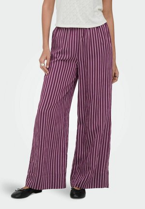 Kvinde iført lilla bukser med vide ben med lodrette hvide striber, sorte ballerinaer og en hvid tekstureret ærmeløs top.