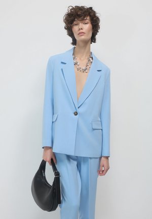 Woman with short curly hair wears light blue suit, layered pearl necklace, and holds a black crescent-shaped handbag against white background.