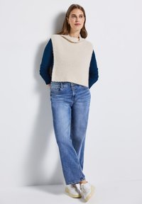 Pull court beige clair avec un col chauve-souris, associé à un jean bleu clair à coupe décontractée et des baskets blanches à semelle légèrement compensée.
