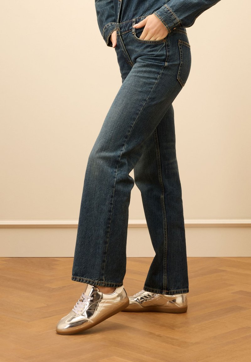 Dunkelblaue, ausgeflogene Jeans mit klassischem Schnitt, kombiniert mit glänzenden, silbernen Low-Top-Sneakern und weißen Schnürsenkeln auf einem Holzfußboden.