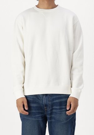 Osoba ubrana w gładką białą bluzę z długim rękawem i niebieskie dżinsy, stojąca na białym tle z rękami swobodnie opuszczonymi wzdłuż ciała.