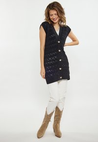 Vest tricotat bleu închis cu un model texturat, având butoane mari din lemn pe față, purtat cu pantaloni albi și cizme de cowboy bej.