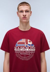 Bordová bavlněná trička s kulatým grafickým motivem zobrazujícím vlajky, "NAPAPURI ANTARCTIC RESEARCH PROGRAM," a vytištěnými GPS souřadnicemi v bílé barvě.