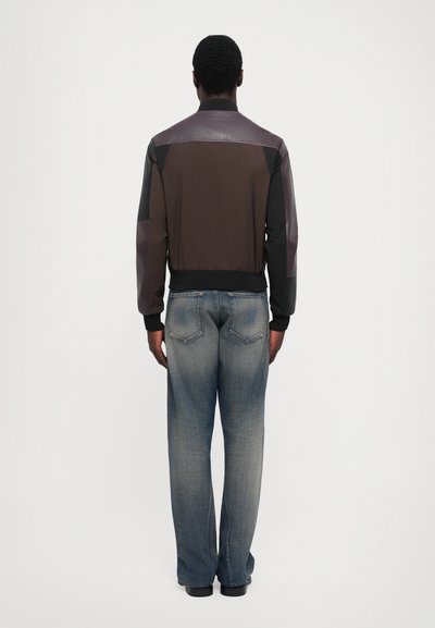 Herrarnas bomberjacka i brunt, med en design i blandade material som kombinerar läder och tyg. Matchas med löst sittande blå denimjeans.