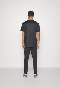 Melnā sporta T-kreklā ar teksturētu apdari, apvienots ar pieguļošām melnām biksēm un pelēkiem sporta apaviem ar sarkaniem akcentiem. Skats no aizmugures.