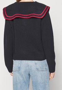 Marinblå stickad tröja med framträdande rödprydd volangkanterad krage. Ribbad nederkant och muddar, kombinerat med ljusblå jeans i denim.