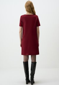 Bordeauxfarbenes Kurzarmkleid mit geradem Schnitt, aus glattem Stoff gefertigt, mit einer Naht hinten und kombiniert mit hohen schwarzen Stiefeln.