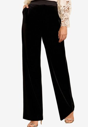 Pantaloni a gamba larga in velluto nero con una texture liscia, caratterizzati da un'alta vita e tasche laterali, abbinati a un top in pizzo leggero.