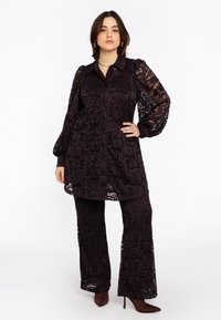 Vrouw met donker haar, gekleed in een bijpassende zwarte kanten tuniek en uitlopende broek, staande met één hand op de heup en bruine hakken.