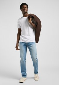 Camiseta blanca de manga corta, jeans rectos de color azul claro y zapatillas beige; sosteniendo un cárdigan marrón texturizado sobre un hombro.