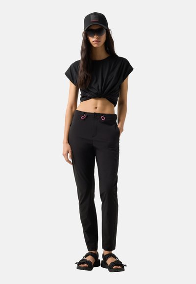 Frau mit schwarzer Mütze, Sonnenbrille, geknotetem schwarzem Crop-Top, schwarzer Hose mit rosa Kordelzügen und schwarzen Schnallensandalen, stehend mit der Hand in der Tasche.