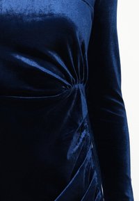 Námořnická modrá sametová šaty s dlouhými rukávy, s detailným zkrouceným uzlem v pase a řasením po straně pro texturu.
