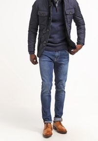 Mansoutfit med en marinblå vadderad jacka, mörkblå tröja, ljusblå jeans och bruna läderskor med snörning.