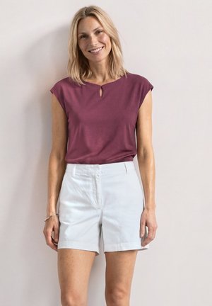 Femme souriante aux cheveux blonds mi-longs, portant un haut bordeaux à manches courtes et un short blanc, debout devant un fond uni clair.