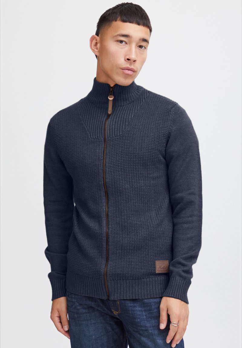 Sweat à fermeture éclair bleu marine foncé avec un motif de tricot texturé, col haut et accent de fermeture éclair marron. Doté d'un écusson logo près de l'ourlet.