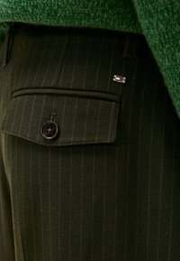 Pantaloni a righe verde scuro con tasca posteriore abbottonata, piccolo logo in metallo e maglione a maglia verde testurizzata infilato dentro.