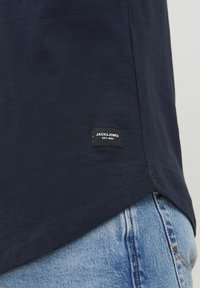 Navy blue cotton t-shirt with a curved hem, featuring a black label with white text near the bottom hem, paired with light blue jeans.