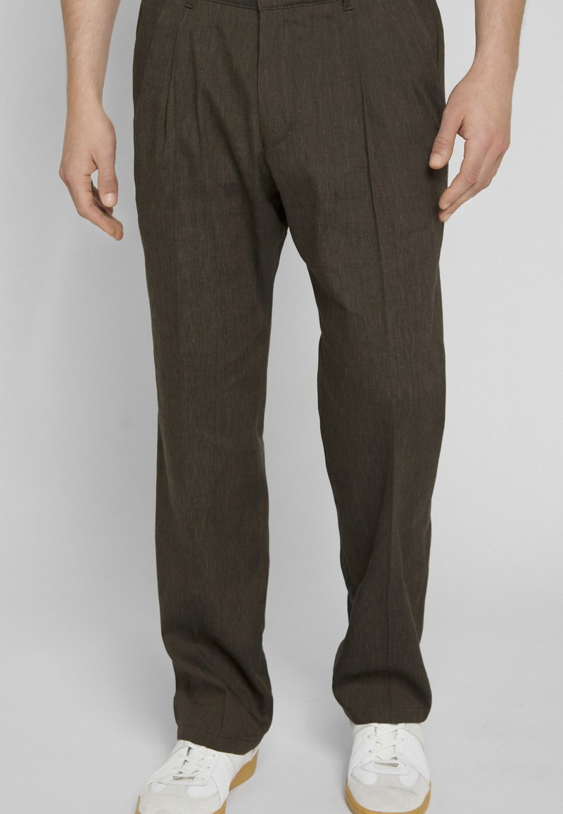 Homme portant un pantalon ample marron foncé et des baskets blanches, debout devant un fond uni gris clair.