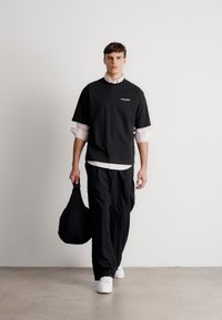 T-shirt preta de algodão sobre uma camisa de botão rosa clara, emparelhada com calças largas pretas e ténis brancos. Segura uma mala preta.