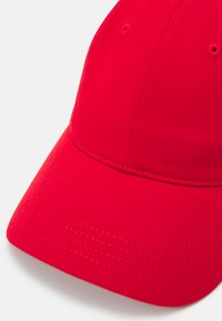 Piros baseball sapka sima textúrával, ívelt emblémával és varrási részletekkel. A korona részén szellőzőlyukak találhatók a légáteresztő képesség érdekében.