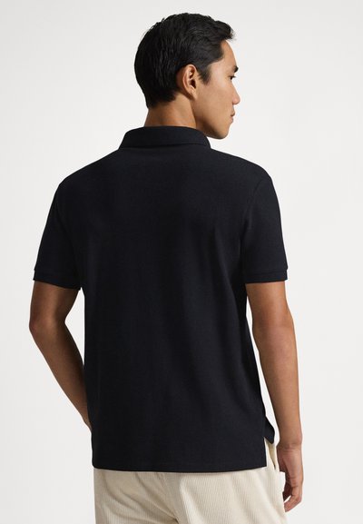 Vyras, trumpais juodais plaukais, dėvintis juodą trumpomis rankovėmis polo marškinėlį ir šviesiai smėlio spalvos kelnes, stovintis nugara į žiūrovą ir žiūrintis į dešinę.