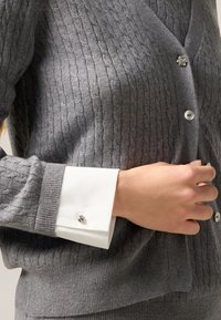 Grau gerippter Pullover mit einem weißen Hemd darunter. Das Hemd hat einen mit Schmuck besetzten Manschettenknopf. Die Knöpfe des Cardigans sind rund und glänzend.