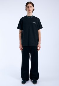 Camiseta de algodón negra con un corte holgado, mangas cortas y un pequeño detalle de logo. Combinada con pantalones anchos negros y zapatos oscuros.