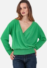 Pull cache-cœur vert avec un décolleté en V profond, manches longues et ourlet côtelé ajusté ; texture douce et coupe décontractée. Associé à un jean bleu foncé.