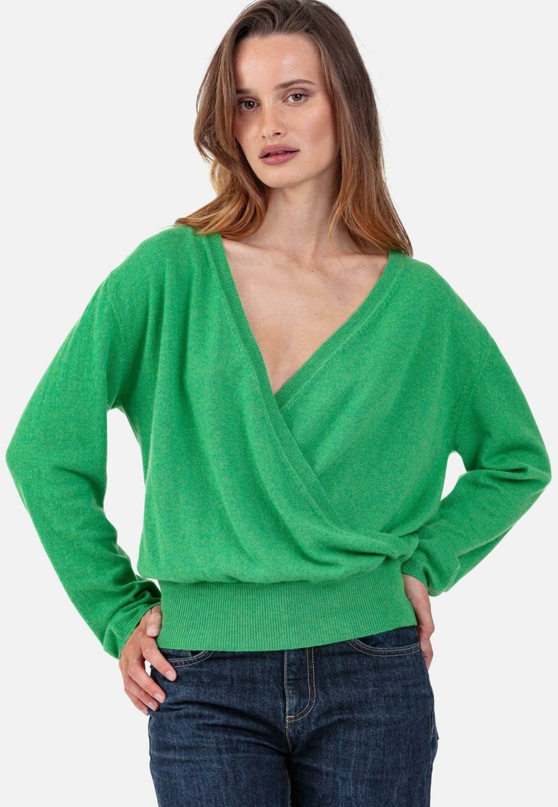 Pull cache-cœur vert avec un décolleté en V profond, manches longues et ourlet côtelé ajusté ; texture douce et coupe décontractée. Associé à un jean bleu foncé.