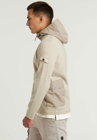 Sudadera de forro polar beige con capucha color marrón claro, detalle de bolsillo lateral y tela texturizada. Presenta una silueta ajustada y acentos en contraste.
