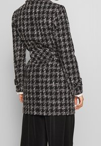 Rutig svart och vit kappa med en strukturerad silhuett, långa ärmar och midjebälte, med texturerat tyg och knappdetaljer.