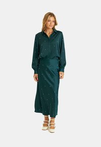 Vestido de satén en color verde azulado con diseño de camisa con cuello, mangas largas y adornos de cuentas blancas dispersos; cuenta con una cintura ajustada y una falda fluida.