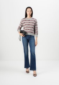 Grauer Pullover mit wiederholtem Textmuster, blaue Schlagjeans und schwarze Handtasche. Das Model trägt schwarze Riemchen-High Heels vor einem weißen Hintergrund.