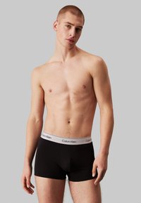 Fekete boxeralsó, széles szürke derékkal, melyen a "Calvin Klein" logó található. Az anyag sima és testhezálló. A modell szürke háttér előtt áll.