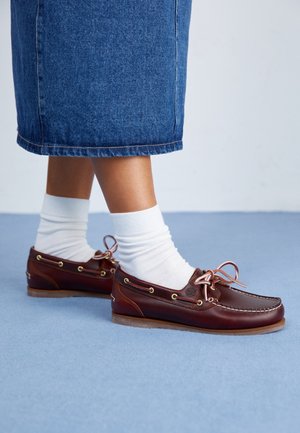 Jambes portant des chaussettes blanches et des mocassins en cuir marron avec des lacets roses, debout sur un sol bleu clair, avec une jupe en jean bleu visible.