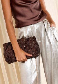 Borsa a mano marrone con paillettes e finitura testurizzata, tenuta in una mano. Pantaloni plissettati in satin argento indossati, caratterizzati da una vestibilità rilassata.