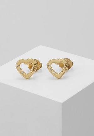 Boucles d'oreilles clous en forme de cœur ton or avec trois petits cristaux et "GUESS" gravé sur chaque, présentées sur une surface blanche.